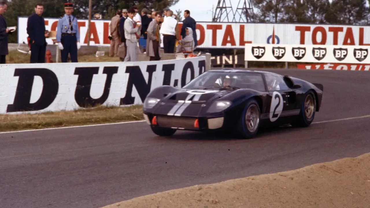Ein schwarzer Ford GT40 Mk3 mit der Startnummer 2 rast auf der Rennstrecke, während Zuschauer und Werbetafeln im Hintergrund zu sehen sind.