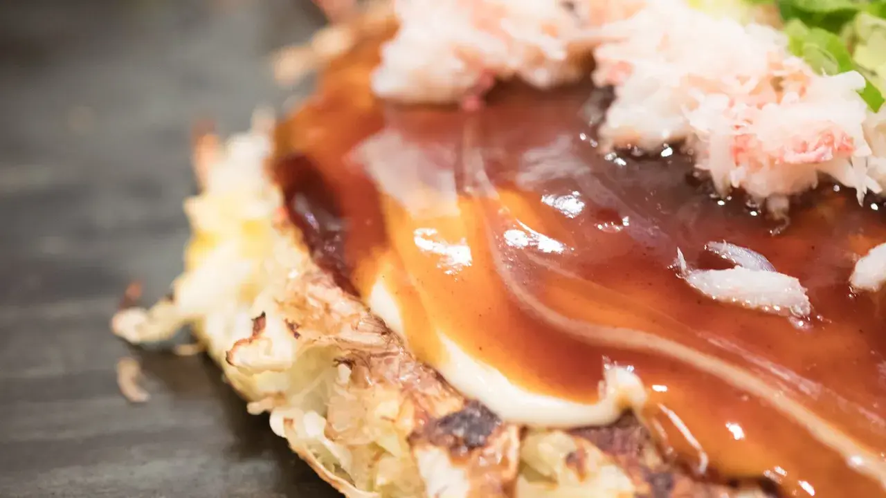 Nahaufnahme eines Okonomiyaki mit cremiger Mayonnaise und würziger Okonomiyaki-Sauce, garniert mit zarten Krabbenfleischstückchen.