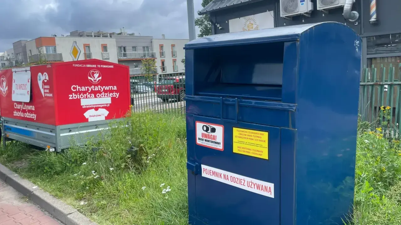 Kontener na odzież używaną obok czerwonego przyczepy z napisem "Charytatywna zbiórka odzieży". To miejsce, gdzie można oddać tekstylia, odpady ubraniowe, zamiast wyrzucać.