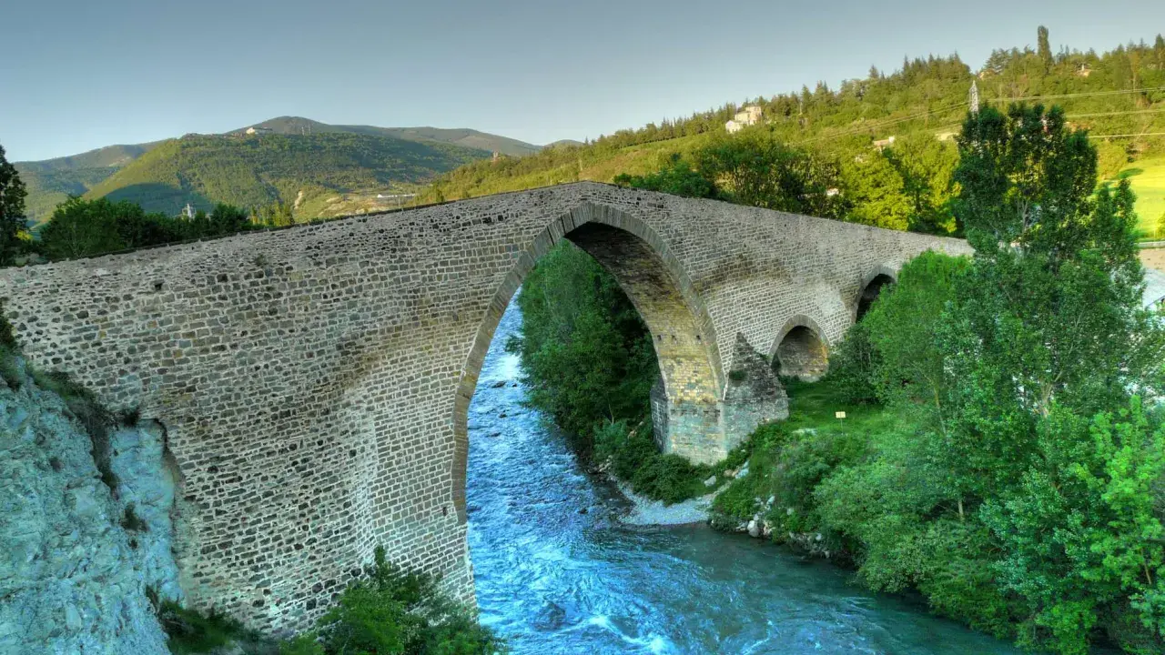 El histórico puente de San Miguel en Jaca cruza un río caudaloso rodeado de vegetación y montañas.