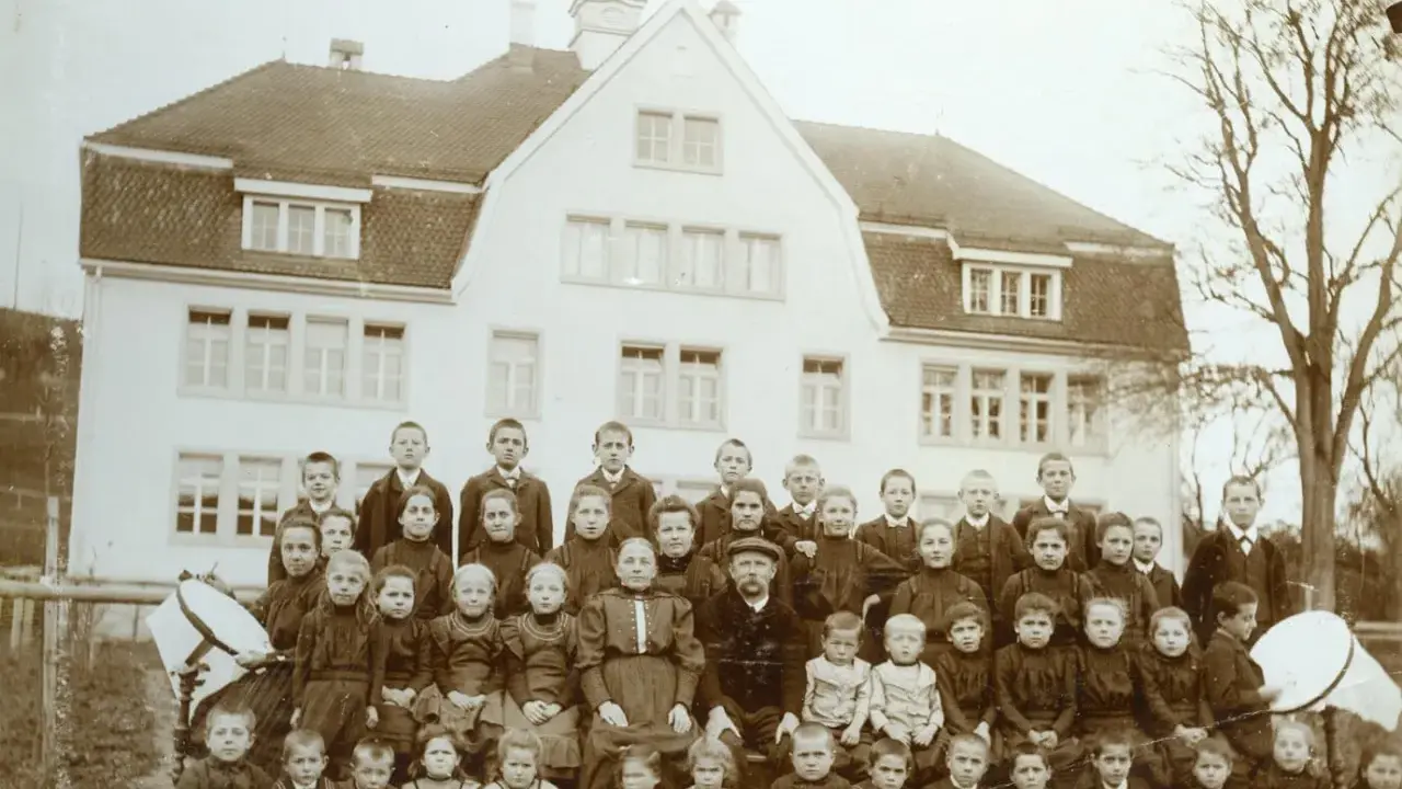 Schulkinder und Lehrer posieren vor einem Gebäude. Die Kleidung deutet auf eine Zeit hin, in der Armut, also Pauperismus, ein großes Thema war.