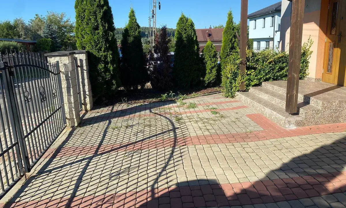 Nowy pomysł na stary chodnik: kostka brukowa w odcieniach beżu i czerwieni tworzy ciekawy wzór przed domem, otoczonym zielenią i ogrodzeniem.
