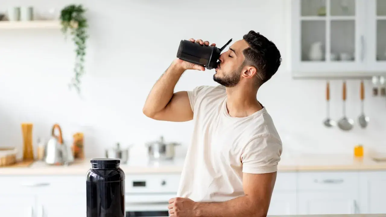 Mann trinkt Proteinshake aus schwarzem Shaker. Daneben steht ein großer Behälter mit Pulver. Wozu Protein**pulver**? Für Muskelaufbau und Erholung.