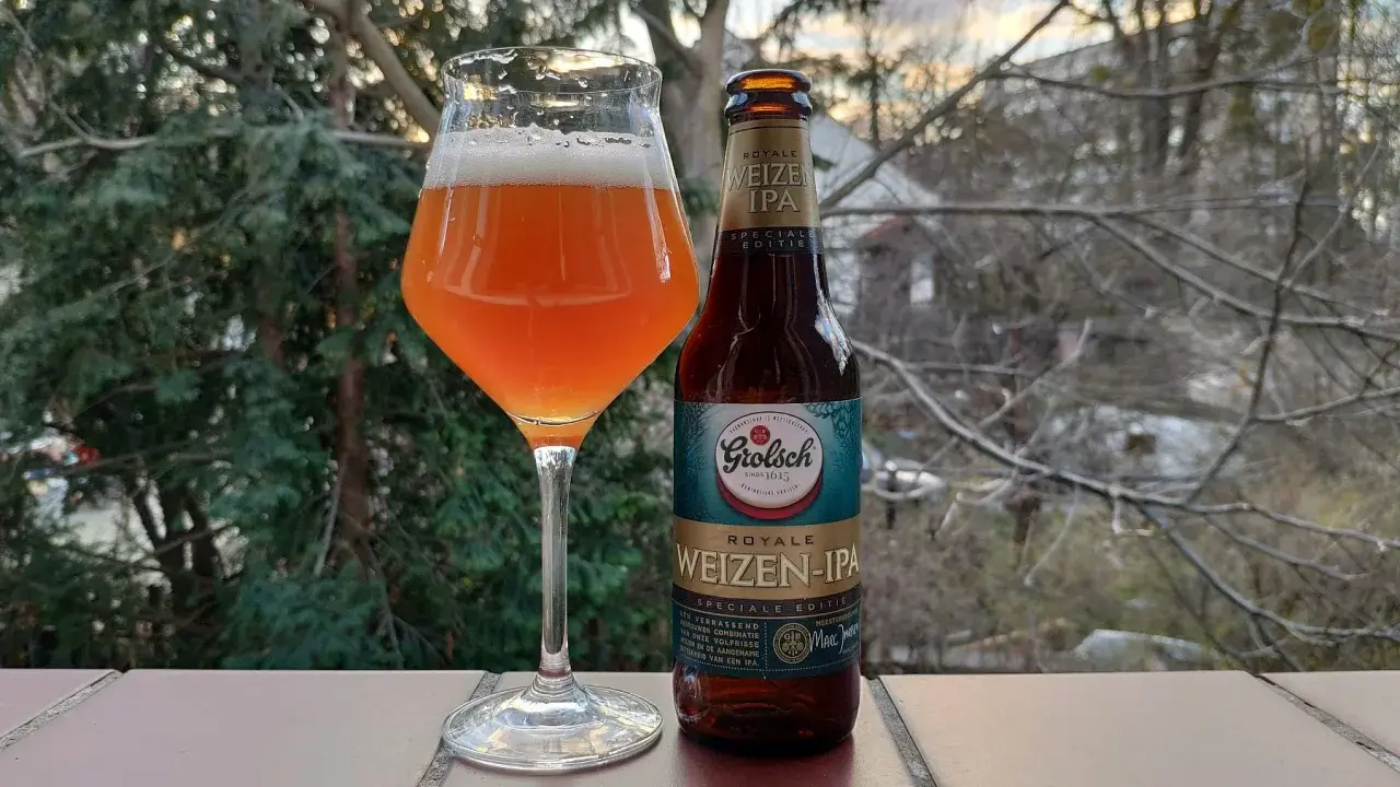 Szklanka z bursztynowym piwem Grolsch Royale Weizen IPA i butelka obok, na tle drzew i nieba.