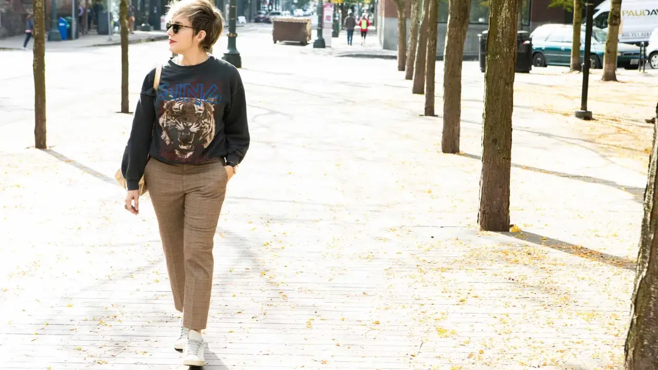 A woman walks down a tree-lined street in a trendy plaid pants outfit, featuring a graphic sweatshirt and white sneakers.