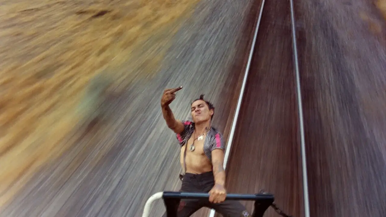 Mike Brodie, un giovane ribelle, fa il gesto del dito medio mentre viaggia su un treno merci, sfidando il mondo con uno sguardo provocatorio.