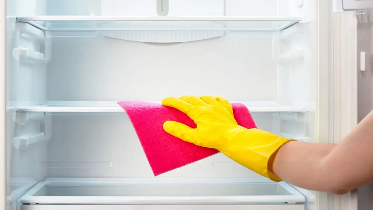 Mano con guante amarillo limpiando estante de frigorífico vacío con paño rosa.