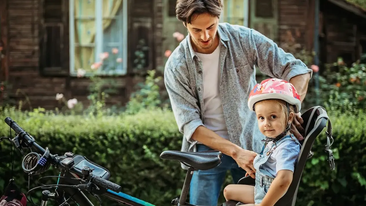 Tata zapina kask dziewczynce, która siedzi w foteliku rowerowym. Od kiedy można wozić dziecko na rowerze? Od kiedy jest bezpiecznie i ma kask!