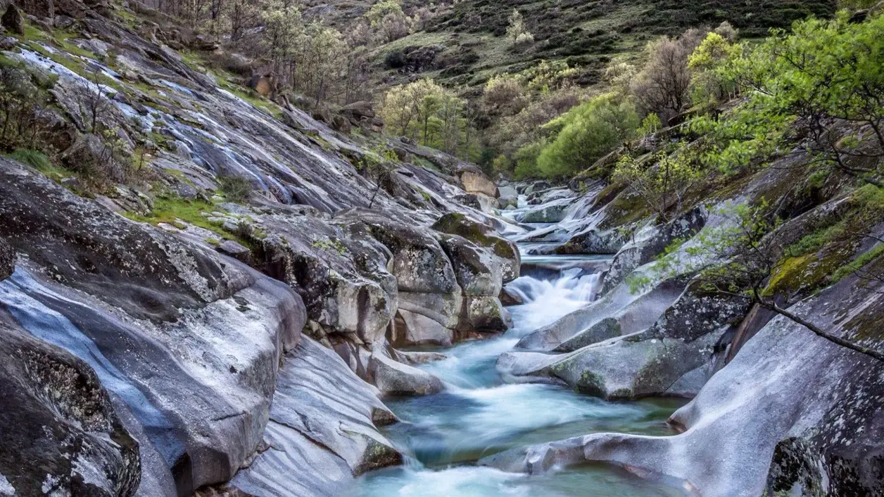 Un río de aguas cristalinas serpentea entre rocas pulidas y vegetación exuberante. Un paisaje idílico que evoca la frescura del verano en Extremadura.