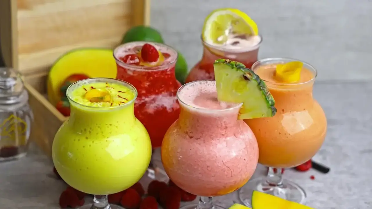 Cinq verres de boisson énergisante maison aux fruits colorés : jaune, rose, rouge et orange, garnie de fruits frais.