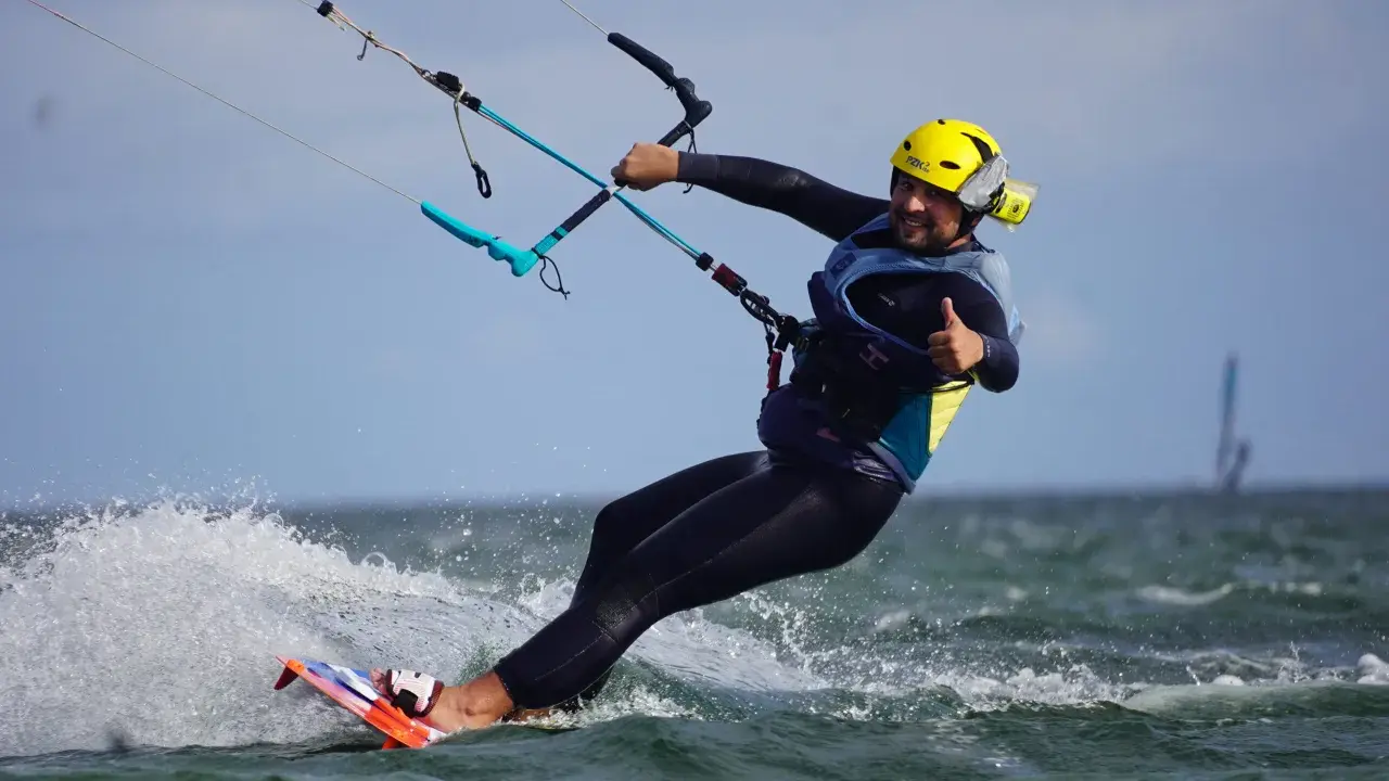 Uśmiechnięty mężczyzna w żółtym kasku i piance cieszy się jazdą na desce. To świetna nauka kitesurfingu!