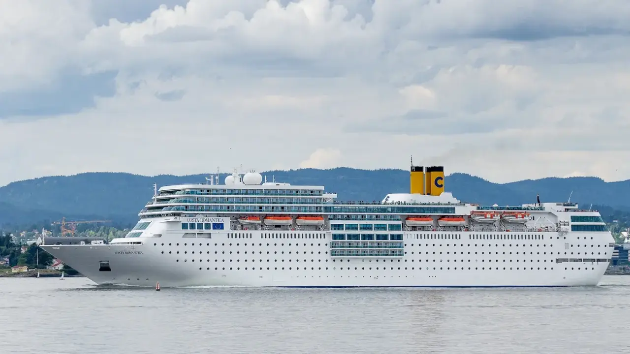 La nave da crociera Costa Romantica naviga in acque calme, con montagne verdi sullo sfondo e un cielo nuvoloso.