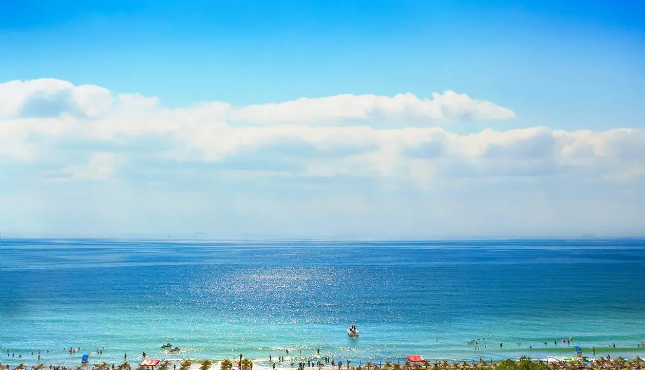 Słoneczny brzeg w pełni lata. Błękitne morze, plaża pełna ludzi i parasoli, a na horyzoncie statki. Idealna pogoda na wypoczynek.