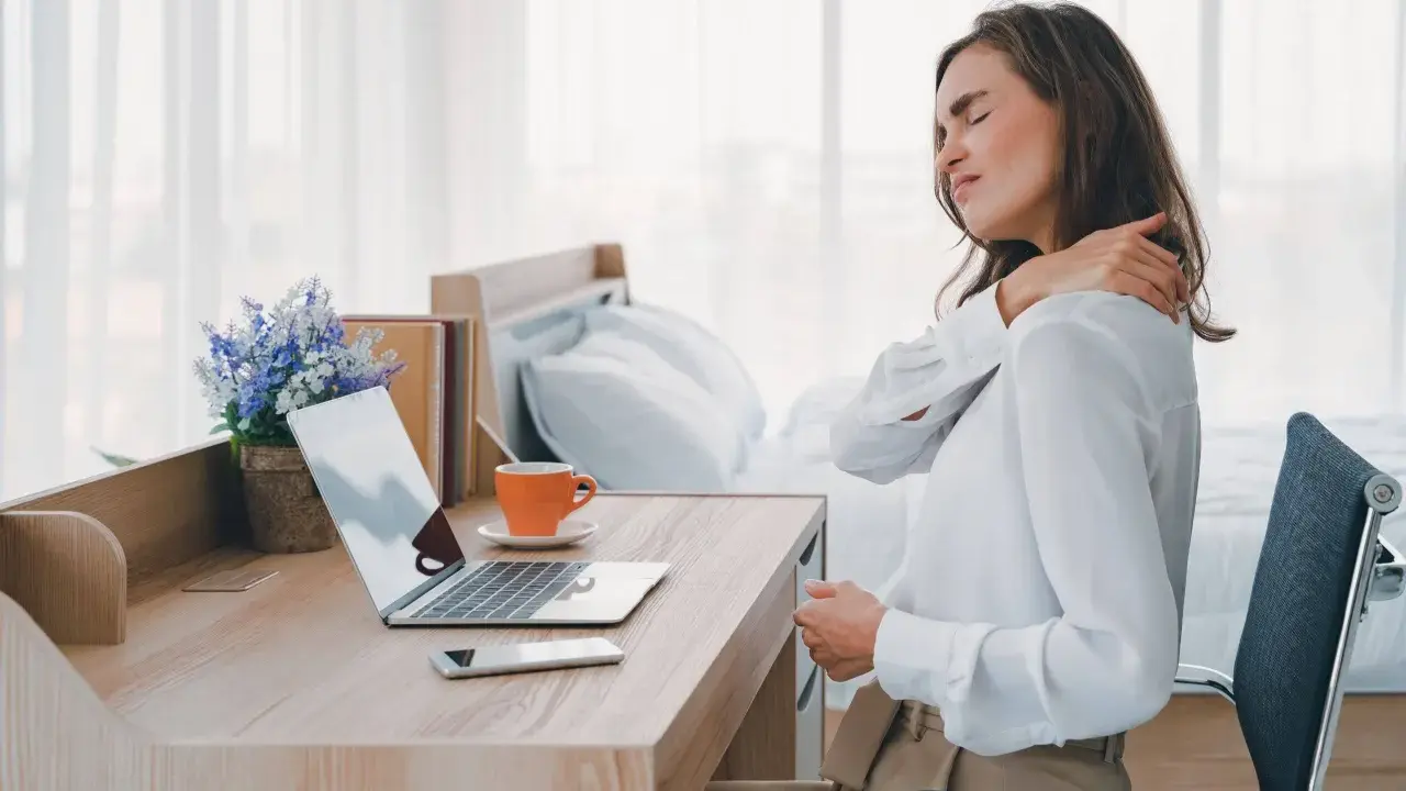Femme se massant l'épaule, assise à son bureau. La mauvaise position bureau peut causer des douleurs.