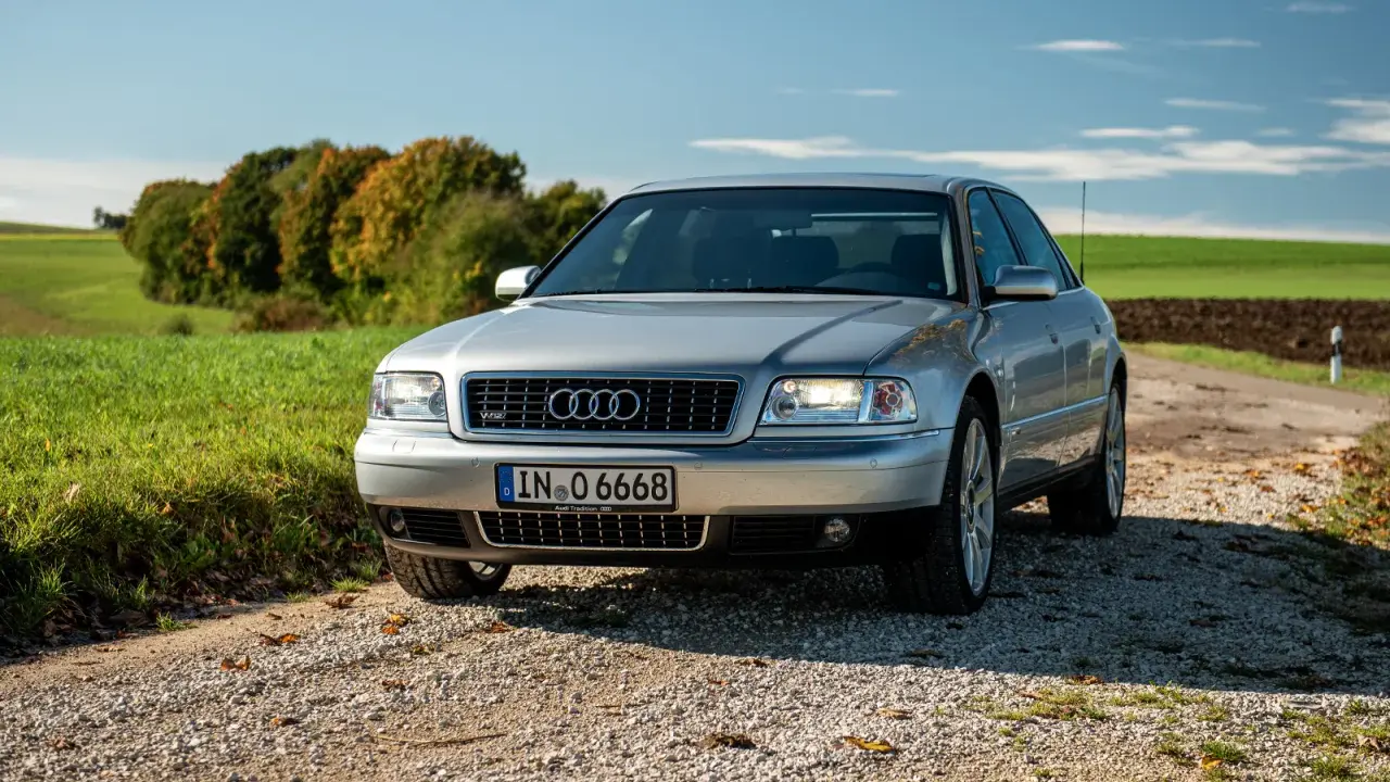 Silberner Audi A8 D2 W12 auf Schotterweg, umgeben von grünen Feldern und Bäumen im Herbstlicht.