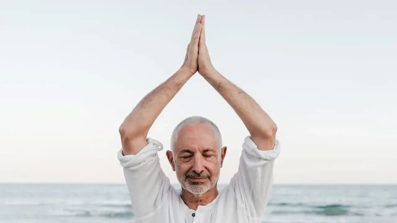 Mężczyzna z zamkniętymi oczami, ręce złożone nad głową, praktykuje mindfulness co to. Spokój nad morzem.