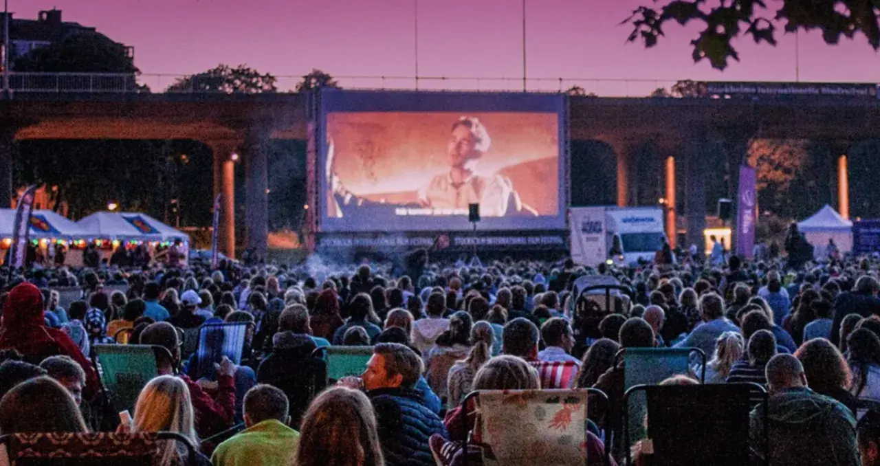 Stor publik njuter av utomhusbio i Stockholm under en rosa kvällshimmel. En stor duk visar en filmscen.