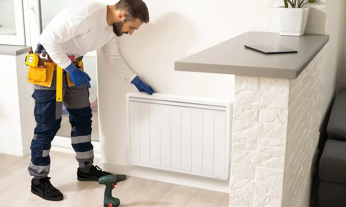Un technicien en bleu, avec une ceinture à outils, installe un radiateur à inertie. Il est prêt à brancher le radiateur inertie sur prise de courant.