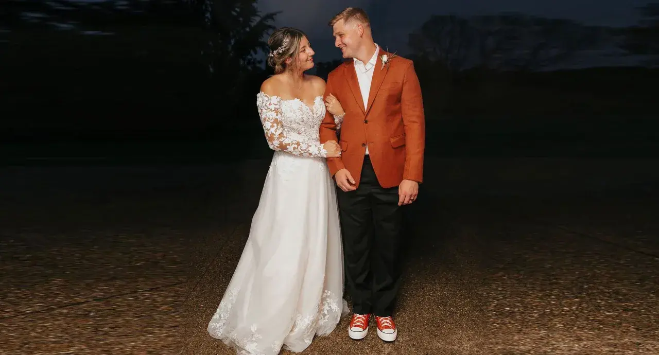 A bride in a lace gown and groom in a rust-colored jacket and black pants, sporting red sneakers, showcase their semi-formal attire for wedding.