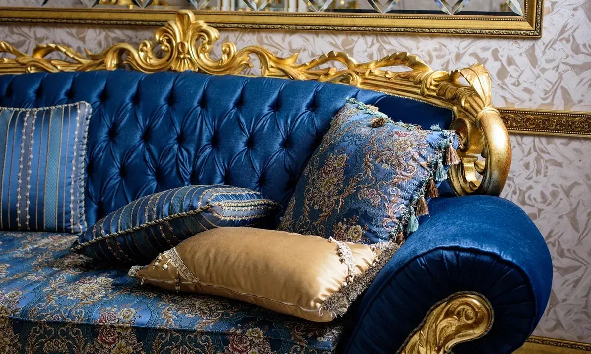 Canapé bleu profond, capitonné, aux ornements dorés, style baroque. Coussins aux motifs floraux et rayés, franges et pompons ajoutent une touche luxueuse.