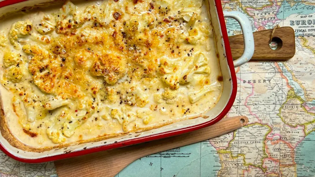Coliflor gratinado con bechamel cremosa, cubierto de queso dorado y pimienta negra, servido en una fuente roja sobre un mapa antiguo.