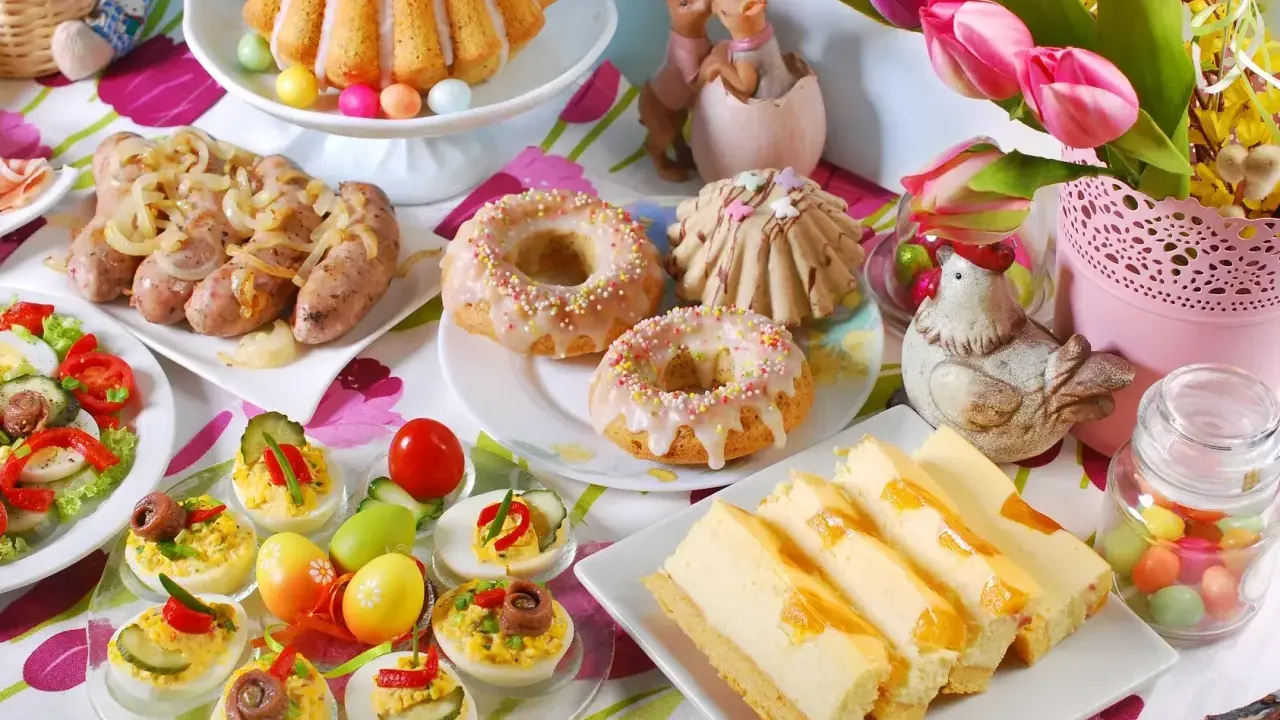 Kolorowy stół z pysznym śniadaniem wielkanocnym: babka, kiełbaski, sałatka, babeczki, sernik i jajka faszerowane.