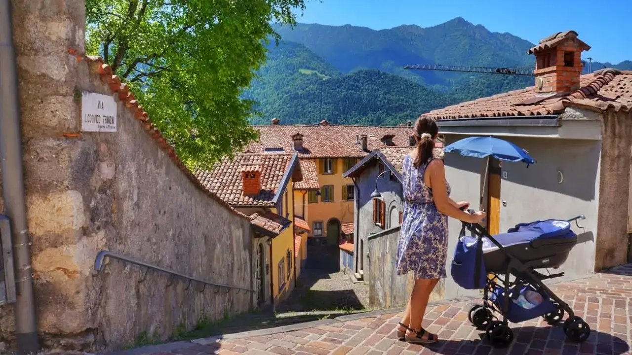 Una donna spinge un passeggino con neonati in montagna, tra case con tetti rossi e un panorama mozzafiato.