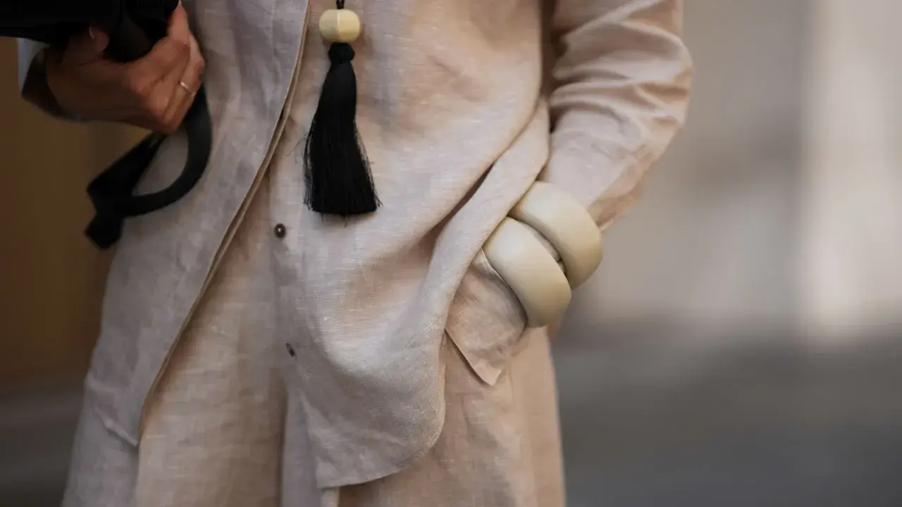 Dettaglio di un outfit in lino materiale, con una mano che tiene una borsa nera, un ciondolo con nappa nera e bracciali color crema.
