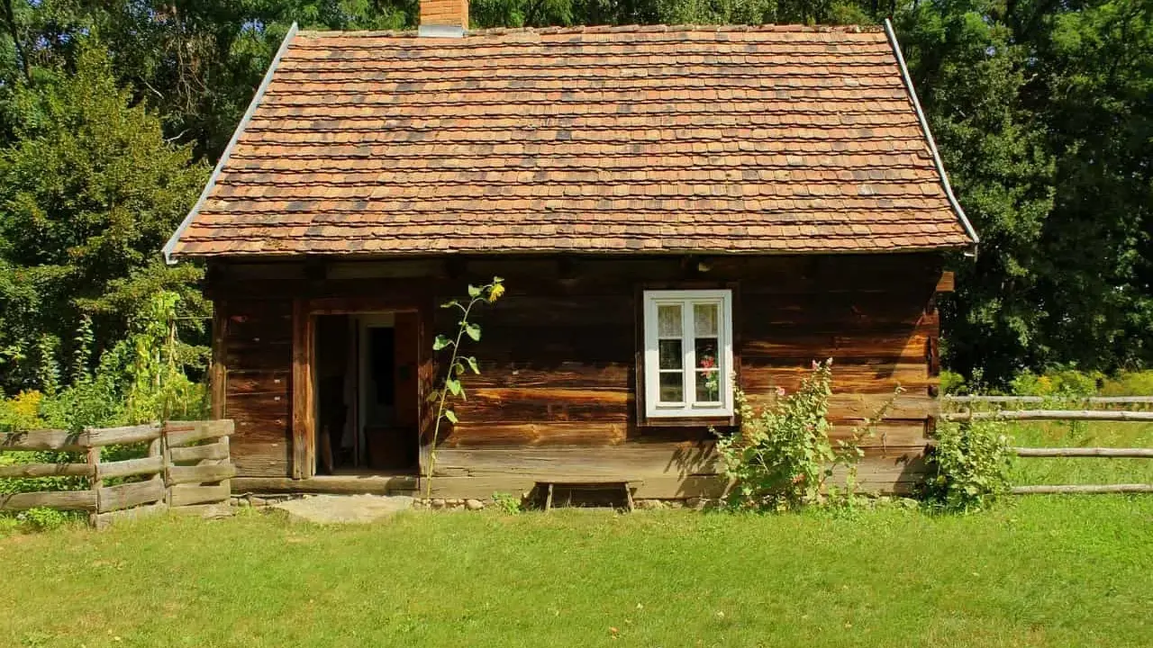 Drewniana chata z glinianym dachem, typowa dla skansenów w Polsce, otoczona zielenią i drewnianym płotem.
