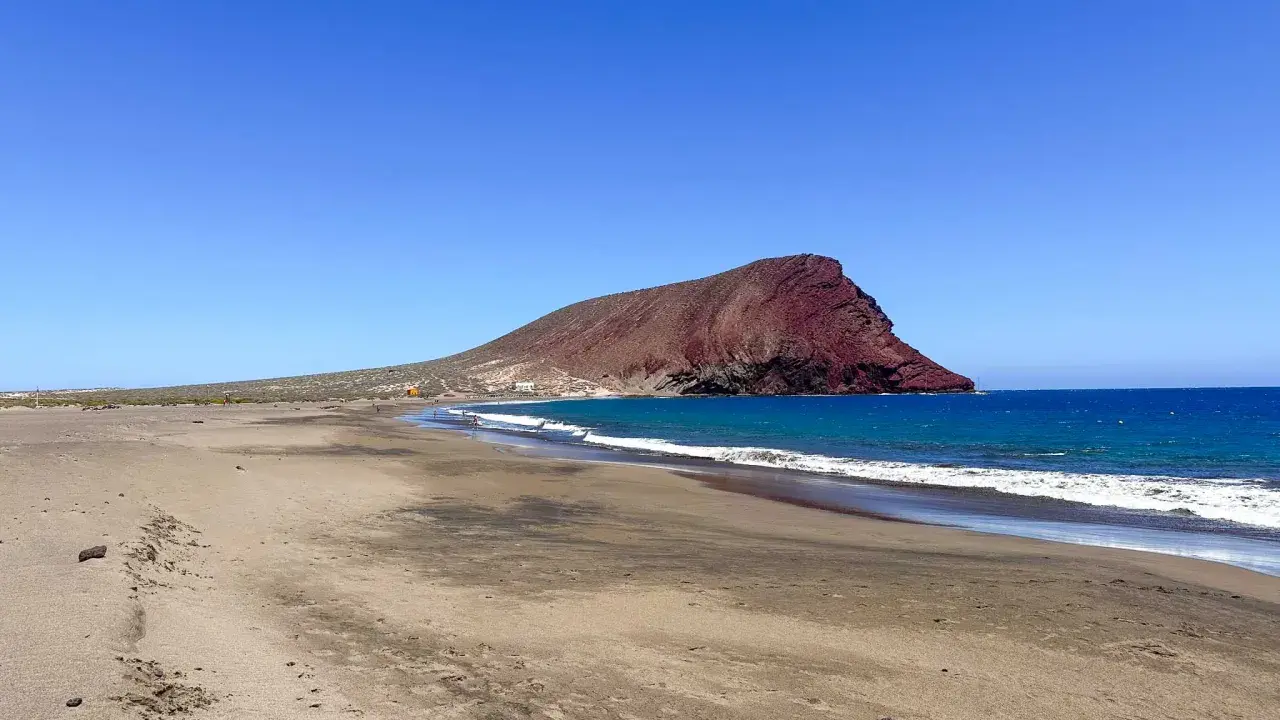 Plaże z białym piaskiem na Teneryfie, z charakterystyczną czerwoną górą w tle i turkusowym oceanem.