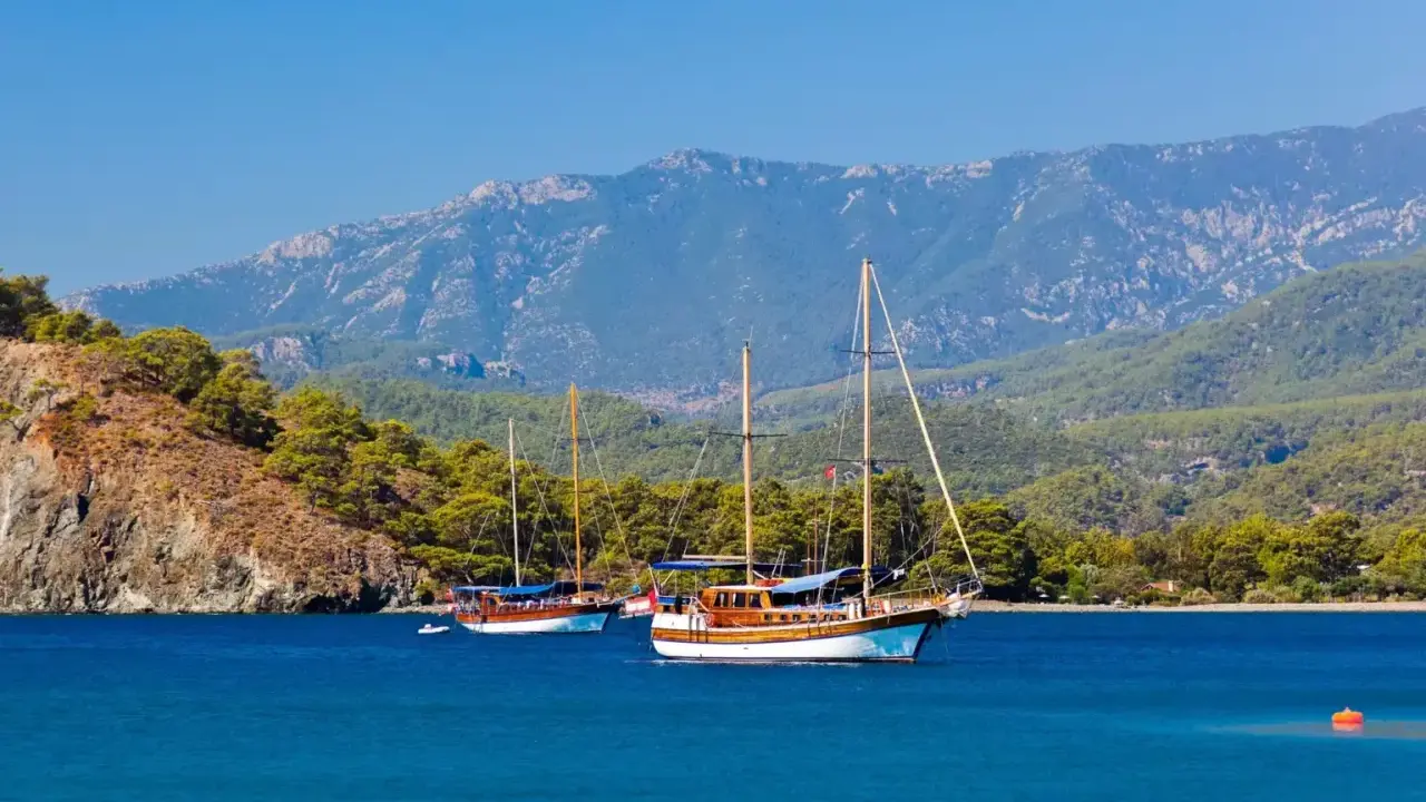 Dwa drewniane jachty kołyszą się na turkusowych wodach, zalesione wzgórza i góry w tle. Idealny dzień na zwiedzanie wybrzeża w okolicach **Antalya miasto**.