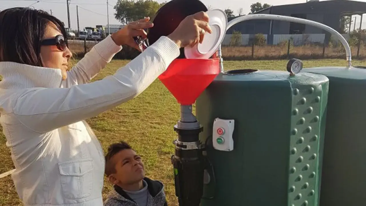 Une femme verse des déchets organiques dans un entonnoir rouge, alimentant un méthaniseur domestique. Un garçon regarde, curieux.