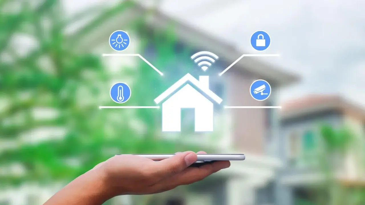 Dłoń trzyma smartfon, z którego wyłania się symbol domu z ikonami oświetlenia, temperatury, zamka i kamery. To wizualizacja systemu zarządzania energią.