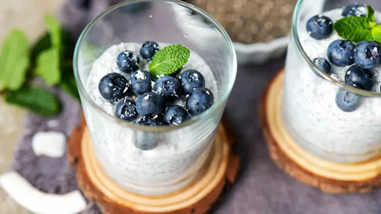 Zwei Gläser mit cremigem Chia Pudding, garniert mit Blaubeeren, Kokosraspeln und Minze. Ein gesunder Genuss!