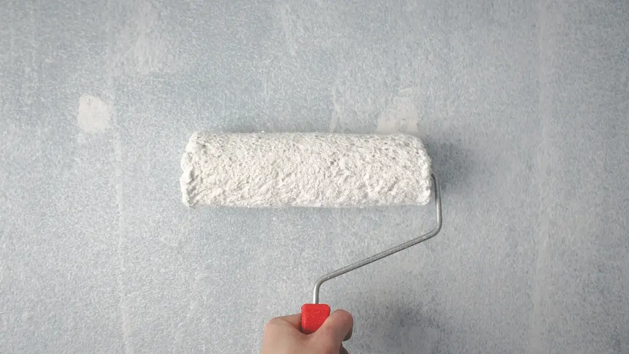 Main tenant, comment rattraper une peinture mal faite. Un rouleau à peinture blanc est tenu par une main sur un mur bleu clair texturé.