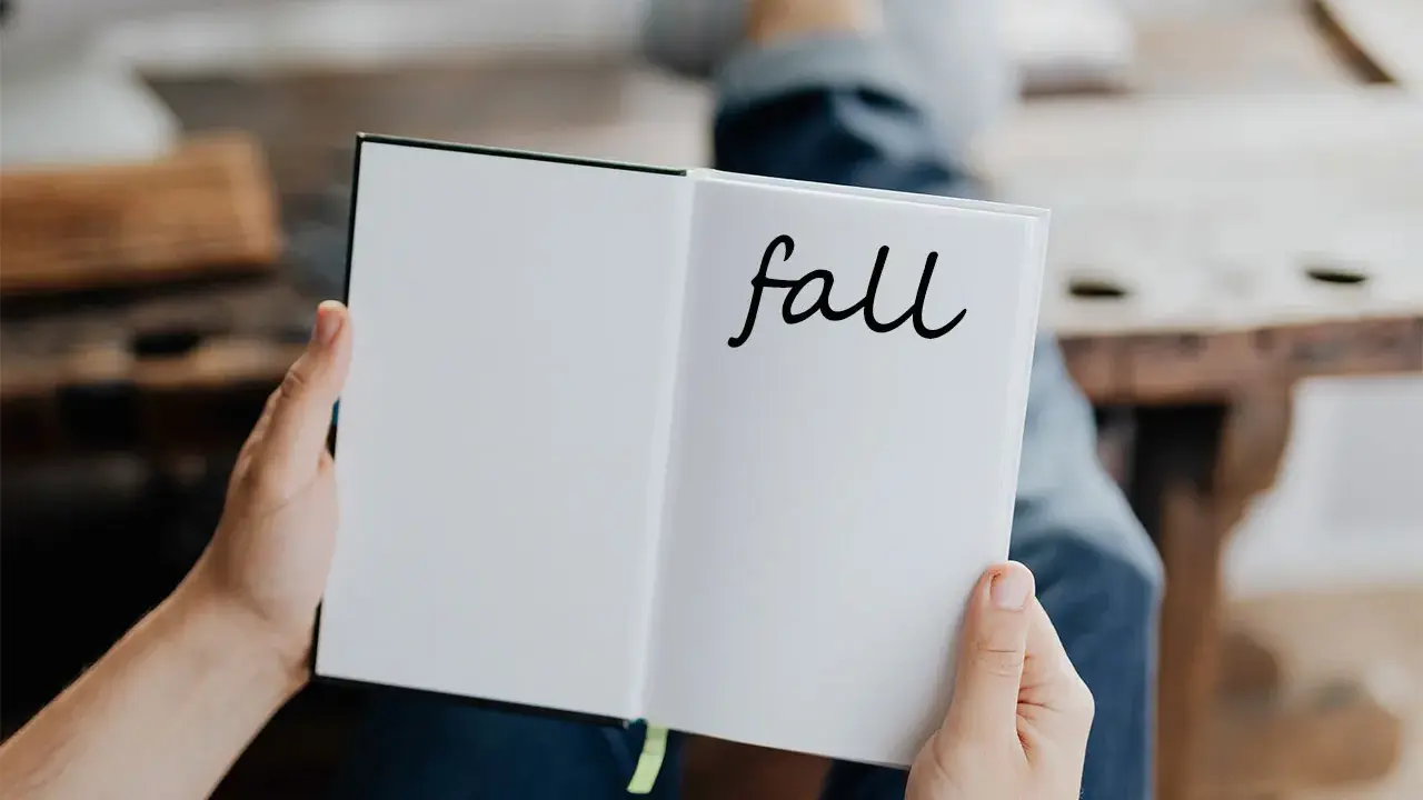 Otwarta książka z napisem "fall" na prawej stronie. Osoba w dżinsach trzyma ją, przygotowując się do jesiennych zapisków.