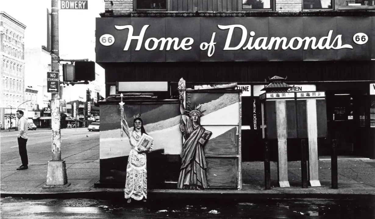 Corky Lee photos: A person in ornate robes stands beside a Statue of Liberty cutout on a wet street, with "Home of Diamonds" sign above.