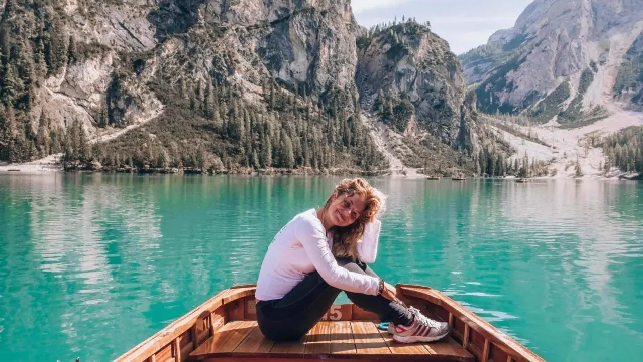 Uśmiechnięta kobieta w łodzi na turkusowym jeziorze w **Włoszech Dolomity**. Majestatyczne góry i lasy tworzą tło dla tej malowniczej sceny.