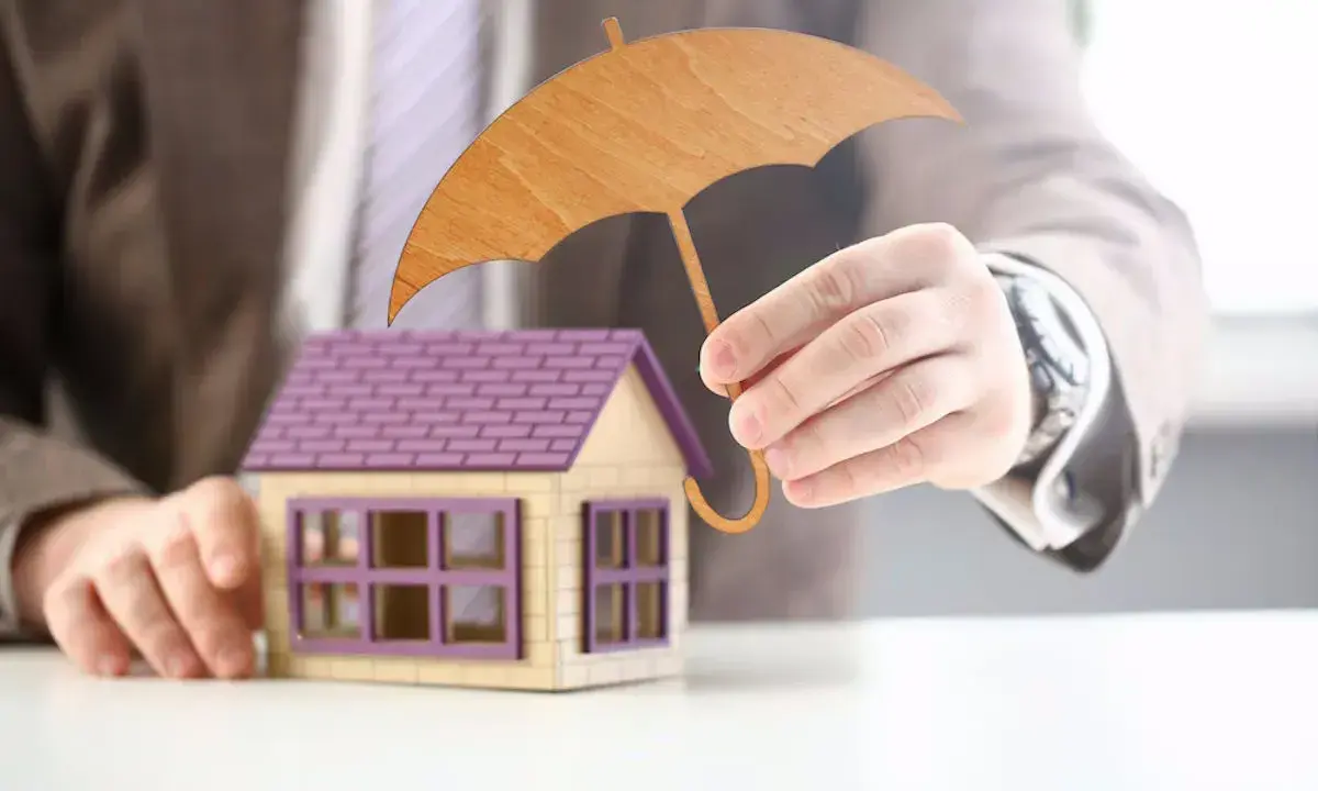 Main d'un homme tenant un parapluie en bois au-dessus d'une maison miniature. Une image symbolisant l'assurance habitation obligatoire.