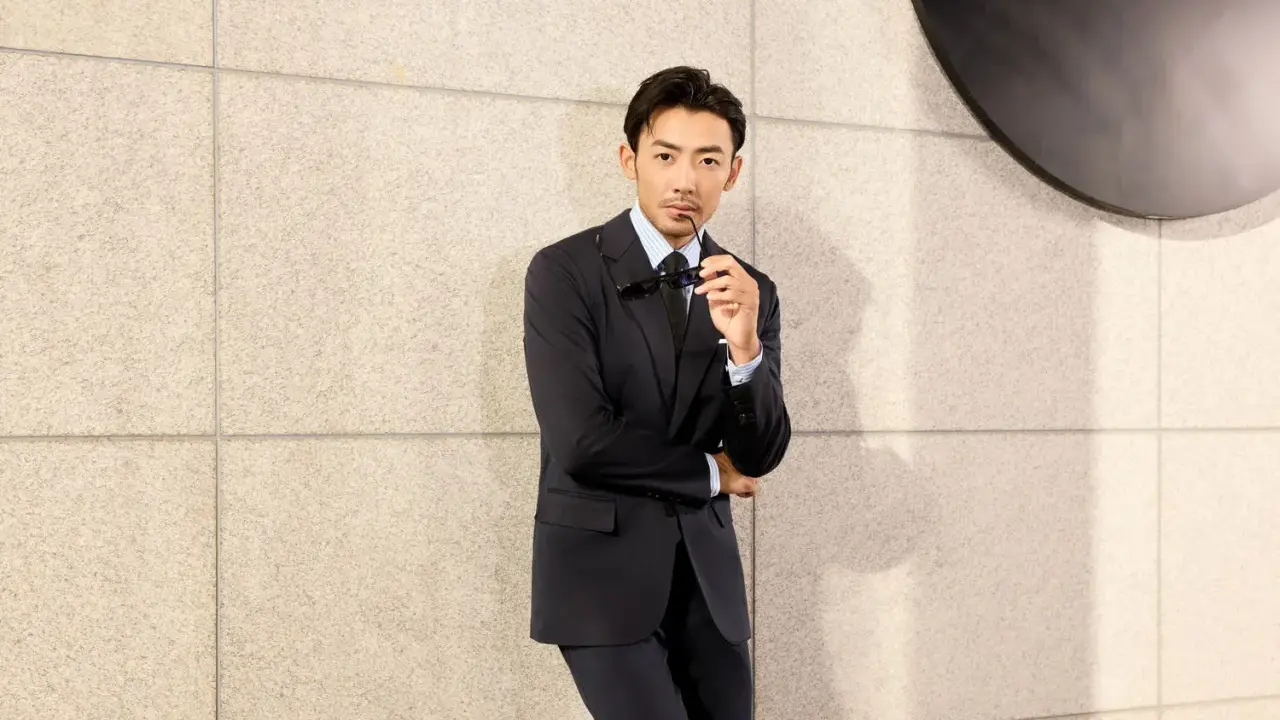 A man in a black suit and black shirt, holding sunglasses, leans against a textured wall.