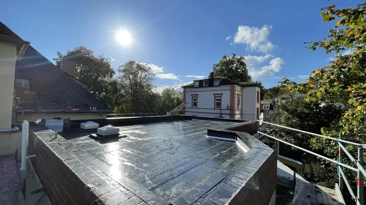 Ein frisch abgedichtetes Flachdach unter strahlender Sonne. Die Dachhaut glänzt, zwei Dachfenster sind sichtbar.