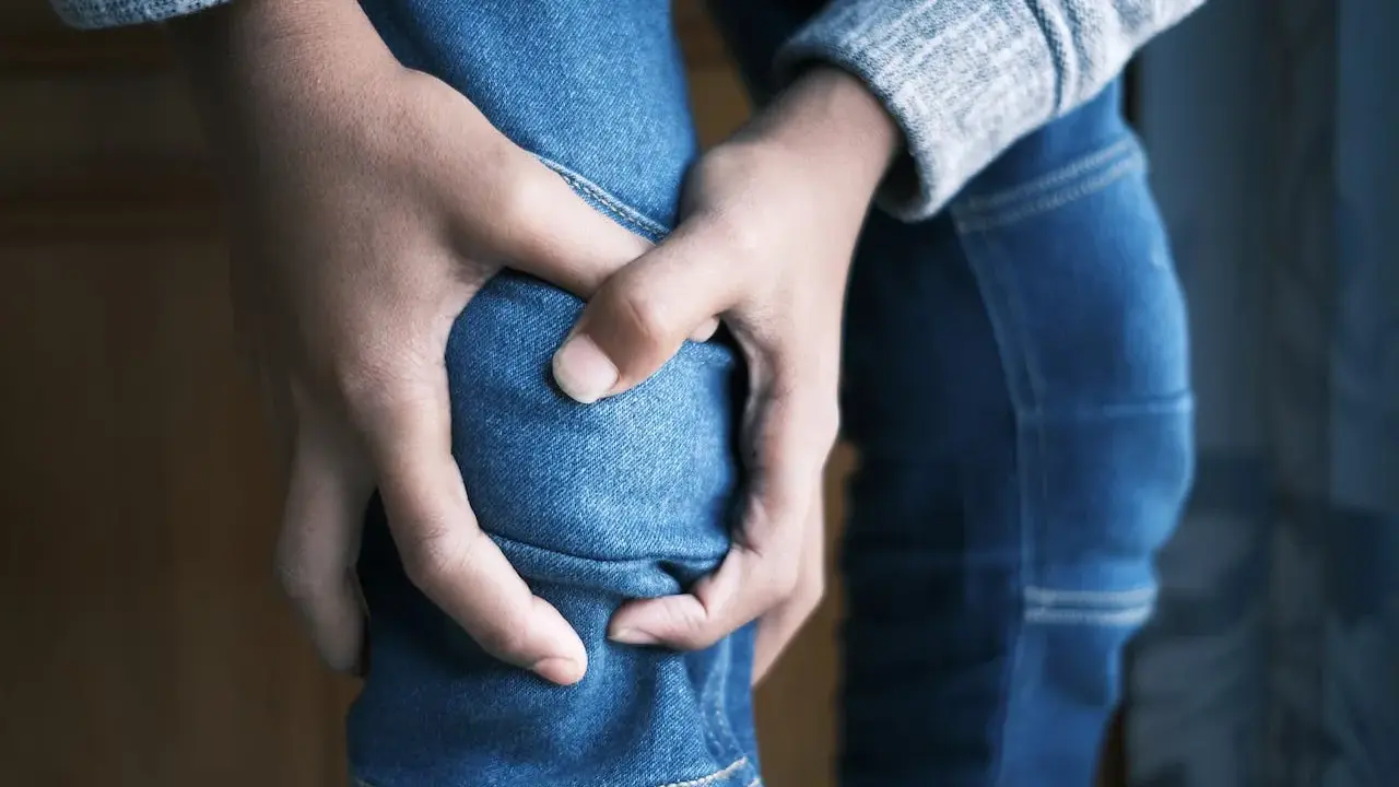 Dłonie zaciskają się na kolanie w dżinsach, symbolizując ból i osłabienie mięśni, być może związane z miopatią alkoholową.