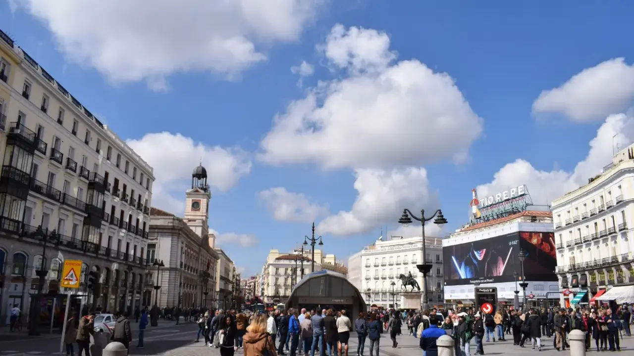 Menschenmassen beleben den Platz Puerta del Sol in Madrid unter einem blauen Himmel mit Wolken. Der Uhrturm und das Tio Pepe Schild sind sichtbar.