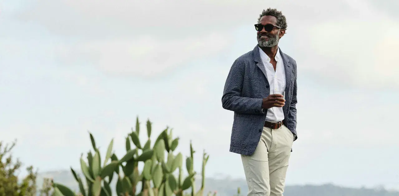 Man in stylish beach wedding attire, holding a drink, with cacti and a hazy sky.