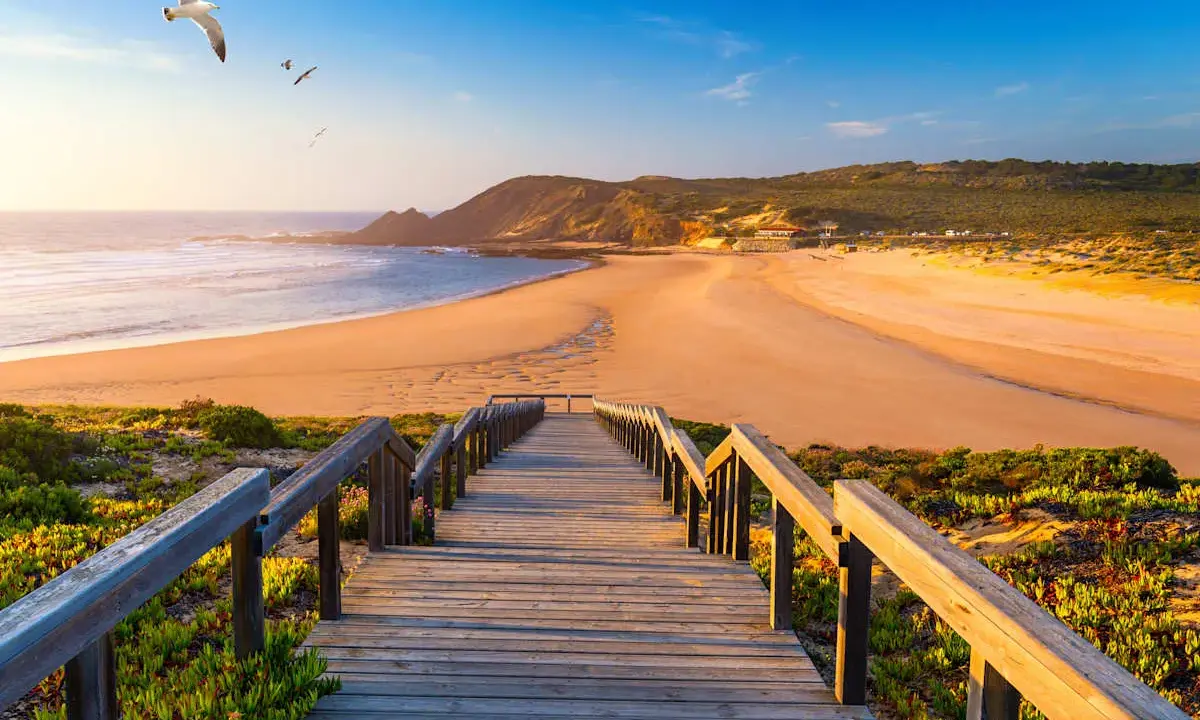 Holzsteg führt zu einem goldenen Strand in Aljezur, Portugal. Möwen kreisen über dem Meer unter blauem Himmel.
