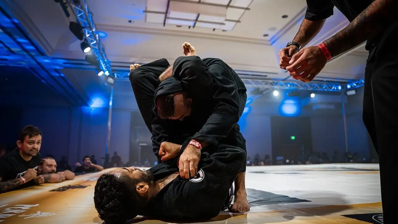 Dwóch mężczyzn w czarnych kimonach walczy w parterze. Intensywna walka jiu jitsu, z widownią w tle.