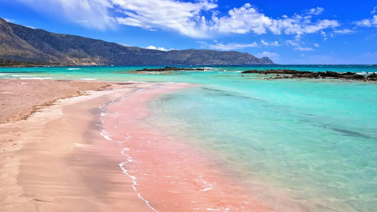 Różowa plaża Kreta kusi turkusową wodą i piaszczystym brzegiem. Góry w tle dodają uroku.