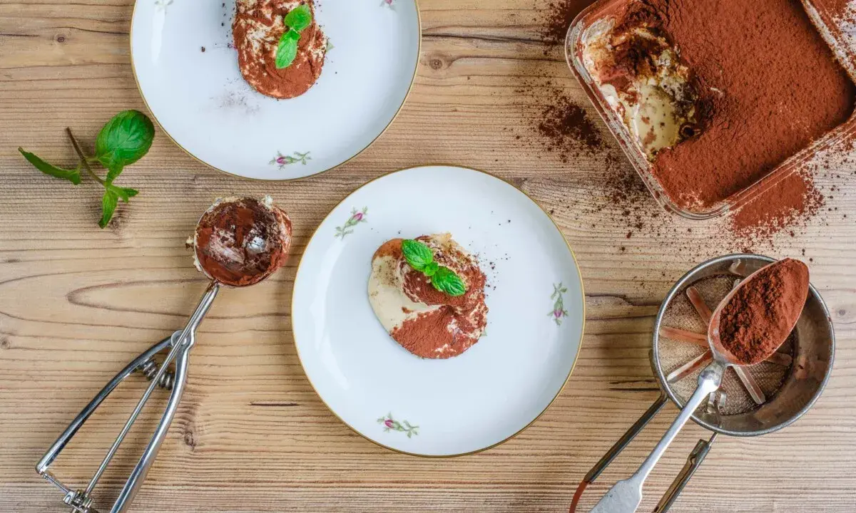 Tiramisu anrichten: Ein Löffel voll cremigen Tiramisus, bestäubt mit Kakao, wartet auf seinen Einsatz. Daneben Teller mit dem fertigen Dessert, garniert mit Minze.