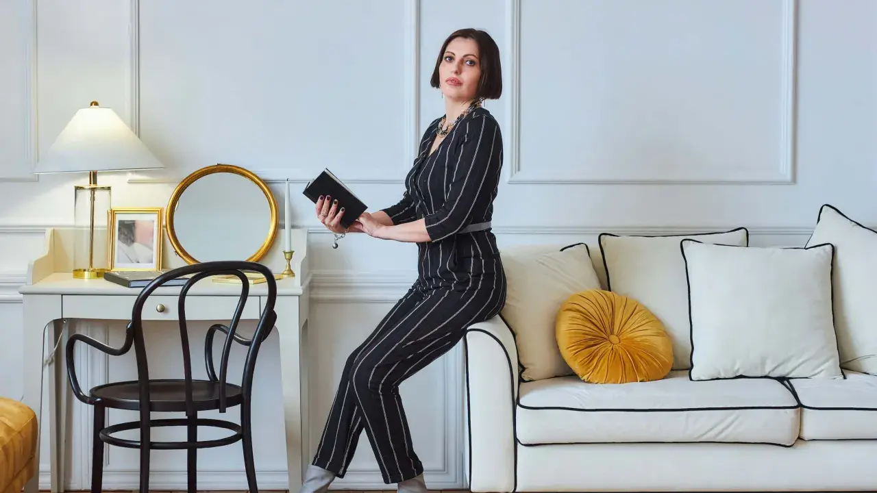 Elegancka kobieta w paski, siedząca na sofie, czyta książkę. Wnętrze z lampą i lustrem to kwintesencja stylu glamour.