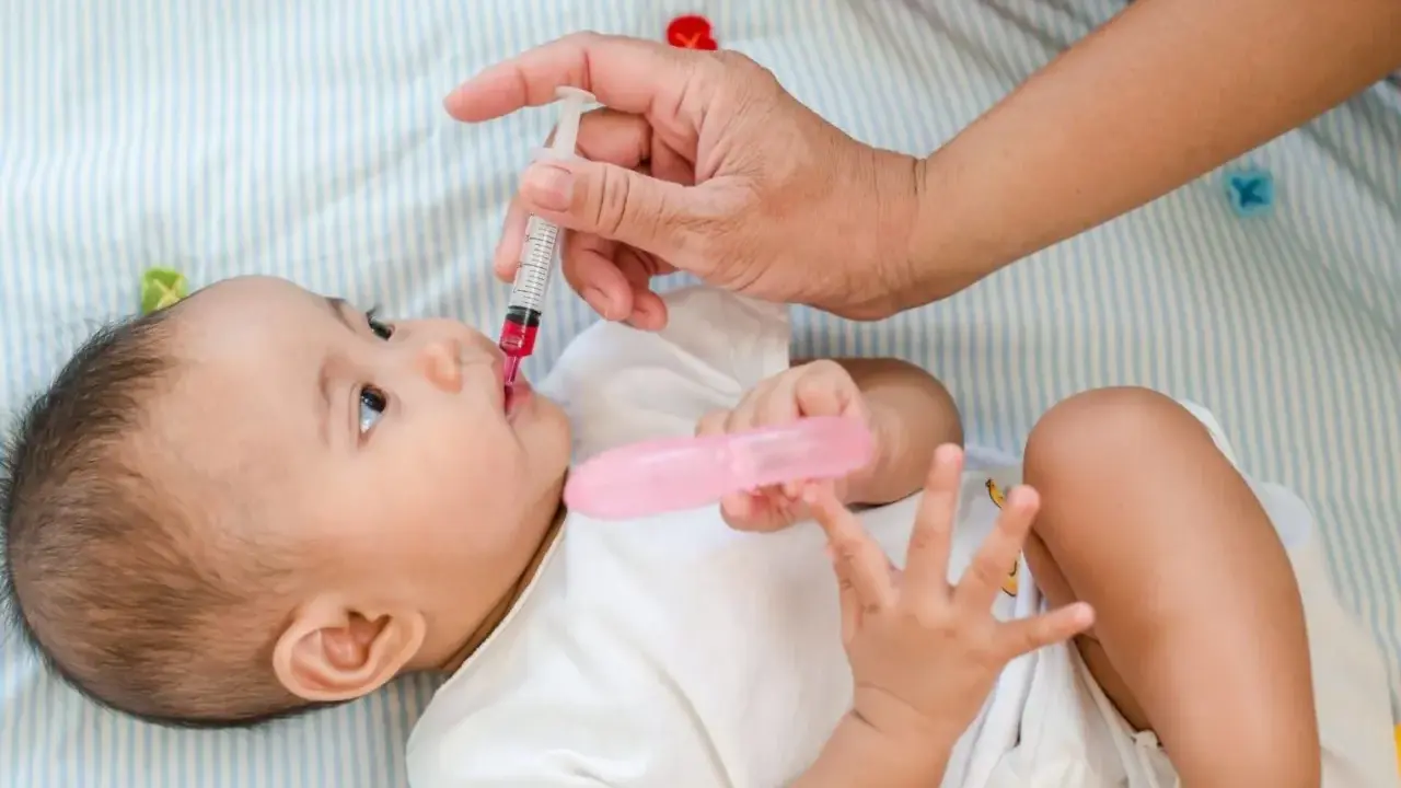 Dłoń podaje niemowlęciu witaminę D z pipetki. Dziecko leży na boku, ubrana w białą koszulkę.